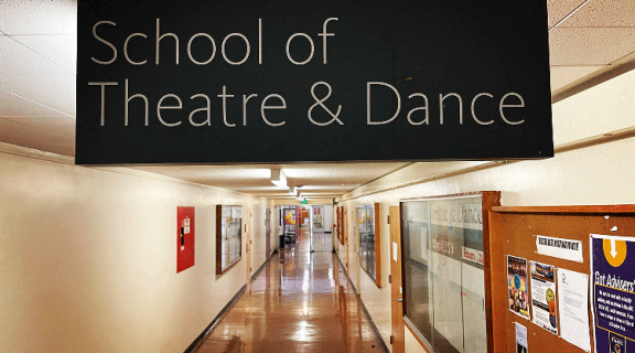 An empty hallway of the Creative Arts Building in which a sign for the School of Theatre & Dance is displayed. The sign is black with white lettering in an elegant, sans serif font.