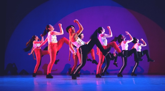 A group of dancers in white tops and red bottoms posing with their right arms and left legs raised in sychronous motion. They are smiling. The lighting is saturated blues and purples.
